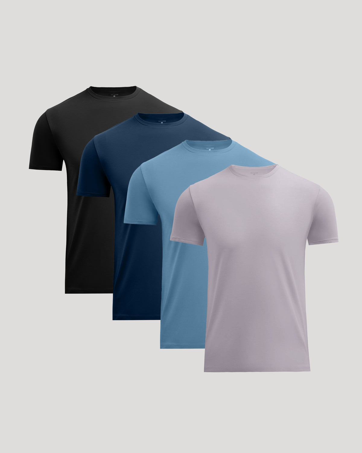 Four short-sleeve crew-neck t-shirts in black, navy, light blue, and dusty lavender, arranged in a staggered diagonal stack on a light gray background.