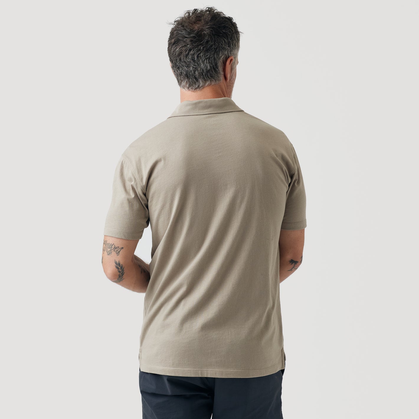 Man in Military Beige Short Sleeve Polo, tattoos on forearms, gray background.