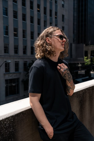 Tattooed Man wearing True Classic Black Crew Neck T-Shirt leaning on a concrete retainer on top of a building