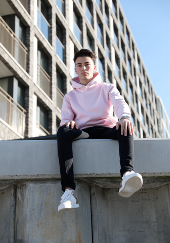 Man sitting on a concreet ledge wearing a pink pullover hoodie and black jeans